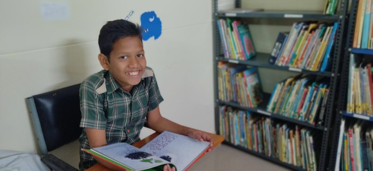 A young boy smiles as he reads and learns about Science at the library at Children of Faith in India
